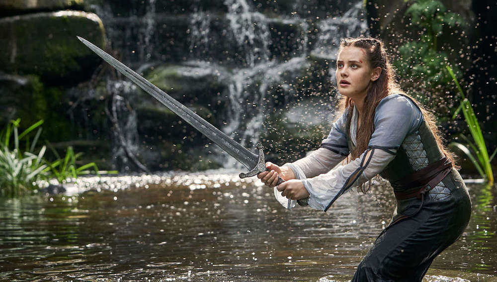 Nimue debout dans une rivière, devant une cascade, l'épée à la main et prête à combattre.