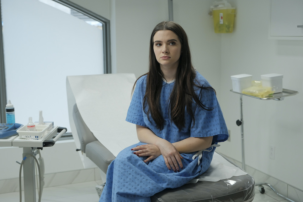Jane assise sur un lit d'hôpital avec une blouse bleue.